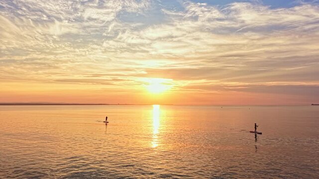 Two people paddle board on calm water as the sun sets in the background. Colors in the sky blend from orange to blue while the sun reflects on the water surface.