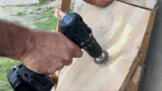 Close-up view of a carpenter's hands skillfully maneuvering a cordless electric drill to clean and texturize a plywood surface, showcasing woodworking, repair, and diy renovation concepts outdoors