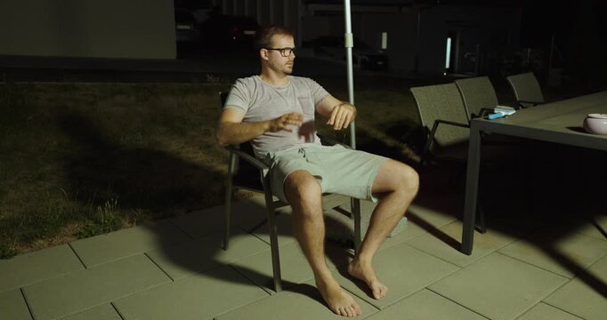 A man sits outside on a patio at night, constantly bothered by mosquito bites and reacting with frustration while trying to enjoy the evening.