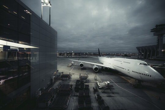 Wide-angle low-key view of a white four-engine passenger aircraft parked at an airport gate with ground service equipment, reflective terminal walls and dense storm clouds