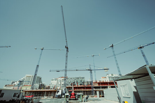 Wide-angle low-angle view of numerous tower cranes rising over an active urban construction site with concrete framework, scaffolding, trucks and clear blue sky