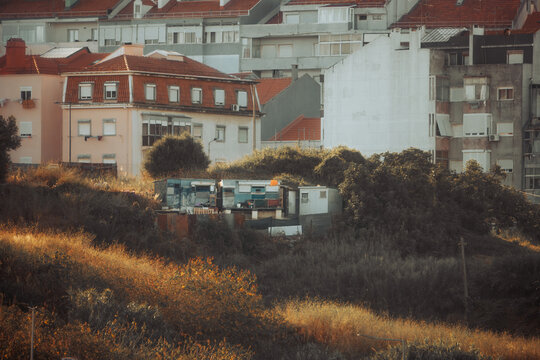 Telephoto view of makeshift shelters clustered on an overgrown vacant lot beneath apartment blocks, revealing urban poverty, housing insecurity and social inequality