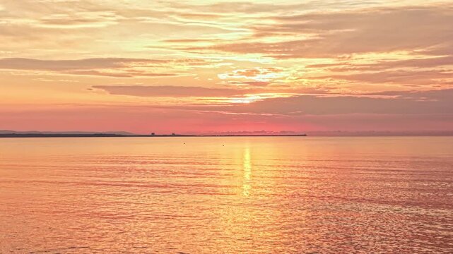 The sun sets over the ocean creating orange and pink colors in the sky. The water reflects these colors, and clouds are scattered in the background. It is evening at the shore.
