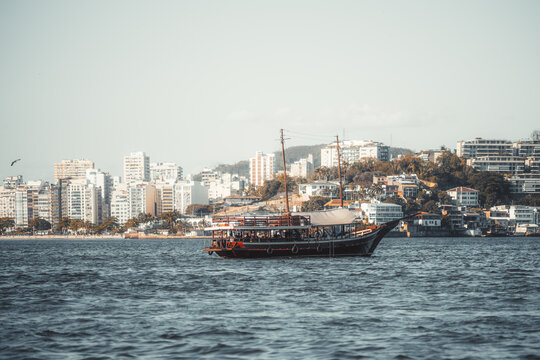 A traditional sightseeing boat with silhouettes of passengers is crossing choppy water, with a dense waterfront skyline and wooded hillside in the background