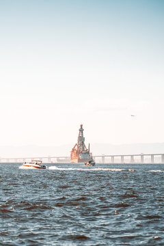 Two recreational motorboats speed across choppy coastal water leaving white wakes, while an offshore drilling vessel, long bridge and hazy hills fill the distance