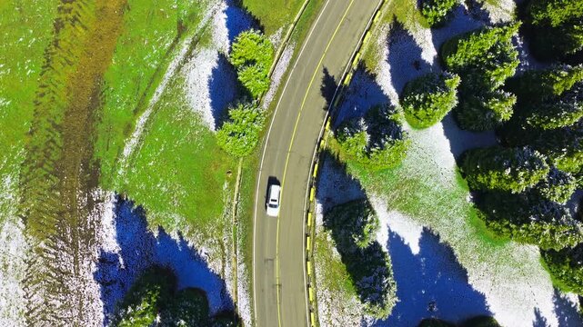Aerial view of a paved mountain road winding through pine trees and light snow cover. High-angle perspective shows contrast between green forest and winter terrain.