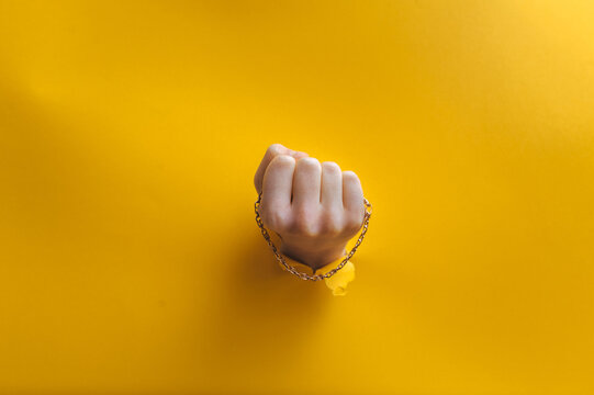 A gold chain in a right child's hand protrudes through a torn hole in yellow paper. Jewelry concept.