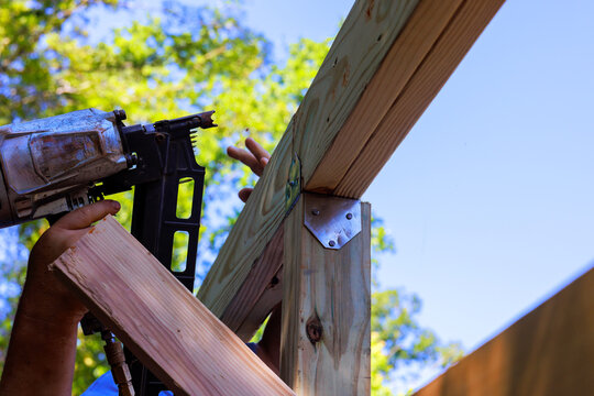 Hands hold air nail gun while securing wooden beams in construction project outdoors