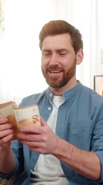 Young man at home holding a stack of euro cash banknotes, counting money with excitement. Caucasian guy on sofa smiles, feeling happy about a big paycheck lottery win or unexpected financial success