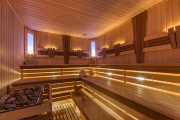 People enjoy relaxation inside a sauna located in a wellness center. Warm wooden benches surround a stone heater. Soft light creates a cozy atmosphere in the evening © Alhim