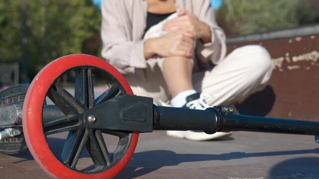 Woman suffering knee pain after scooter accident. Adult woman experiencing severe knee pain after falling off scooter, sitting in skate park and seeking urgent medical