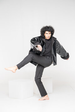 Young man with curly black hair crouching barefoot on white cube, wearing crocodile-embossed leather jacket and beige tweed suit, studio fashion portrait on white background