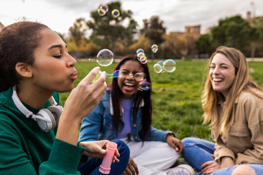 Group of diverse young women sitting in a green park on a sunny day, laughing and blowing soap bubbles while enjoying carefree friendship, summer leisure and joyful connection