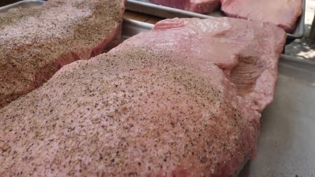 close-up shot of raw beef briskets on a metal tray, seasoned with a dry rub of salt and coarse black pepper. A pitmaster in a black glove prepares the meat for slow smoking in a professional Texas-sty