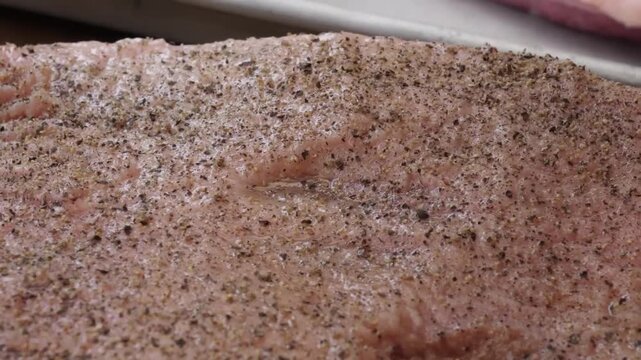 Macro shot of a fresh raw beef steak being prepared for the grill. The footage highlights the detailed marbling of the meat and the coarse texture of freshly cracked black pepper and sea salt seasonin