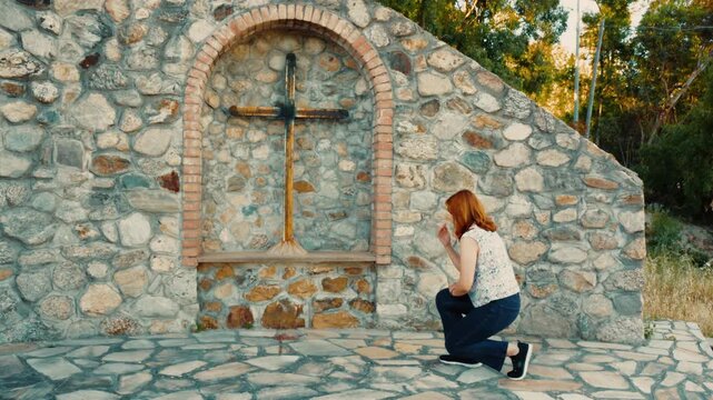 Woman kneels to pray in front of sacred religious crucifix outdoors for peace