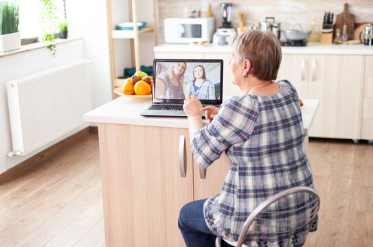 Happy senior woman talks with family by laptop video call in the kitchen, connecting with daughter and niece through online communication, with the action centered on breakfast, technology and a.