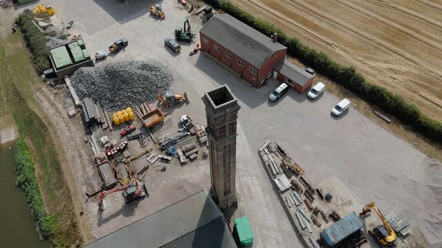 Aerial view of active construction site with heavy machinery. Workers move materials around a central storage area. Stacks of gravel and timber surround the site