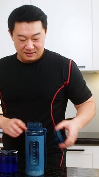 Man adds supplement powder into shaker bottle in kitchen preparing isotonic or creatine drink. Shaking bottle mixes powder with water for workout hydration muscle recovery energy fitness routine gym