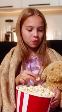 Teenager girl at night home watches cartoon hugs plush toy holds popcorn bowl under warm blanket. Caucasian child kid on sofa at evening smiles chews snack keeps toy close enjoys film time fun mood