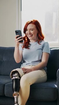 Young woman with prosthetic leg sitting on blue sofa and talking on smartphone, eye level shot