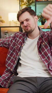 Dislike. Upset unhappy young man showing thumbs down sign gesture, expressing discontent, disapproval dissatisfied bad work at home apartment indoors. Displeased angry Caucasian guy sitting on sofa.