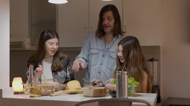 Single father enjoying an affectionate moment with his teenage daughters while cooking together and preparing a meal in the kitchen during evening dinner