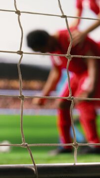 Vertical frame: Soccer player shoots ball into net across the line, with defeated goalkeeper, missed save, selective focus inside goal frame, and blurred footballers.