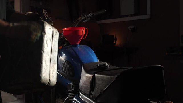 Hand pouring gasoline from a metal jerry can into a classic blue motorcycle's fuel tank using a red funnel inside a dimly lit garage, highlighting maintenance and retro style