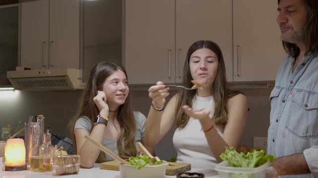 Affectionate father and two teenage daughters enjoying a fun cooking session, laughing and tasting salad together in a cozy home kitchen, showcasing family bonding and collaboration