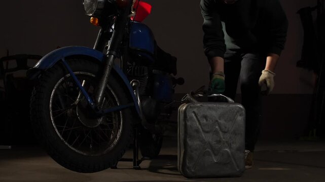 Person standing beside a classic blue motorcycle with a metal jerry can, preparing for a journey or maintenance in a dimly lit garage