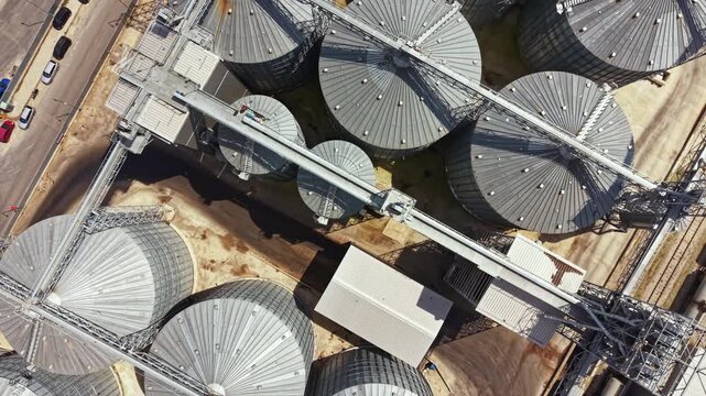 Silos and storage units are seen from above in a rural area. The structures are arranged in rows. The ground shows signs of dirt and dust. Cars are parked along the road nearby.