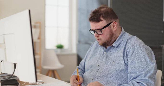 Man accountant in office writing finance analysis. The worker notes figures at the computer, updates budgets, checks reports and records data carefully. Clear theme of focused financial work.