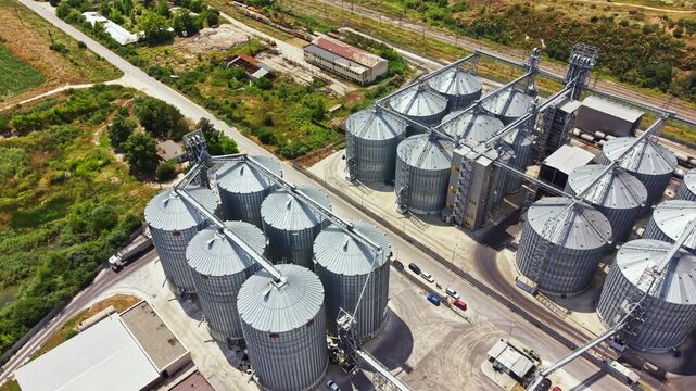 Multiple tall grain silos stand in a rural area with a railway track nearby. Green fields surround the facility, and vehicles can be seen near the entrance.