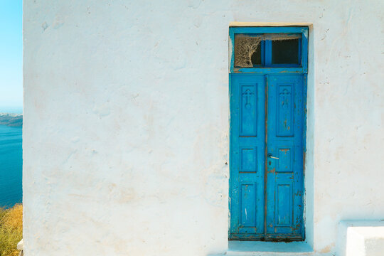 Bright blue door set in a whitewashed wall with a small cobweb above, overlooking the Aegean Sea. The vivid color and aged texture evoke timeless Mediterranean simplicity and coastal serenity.