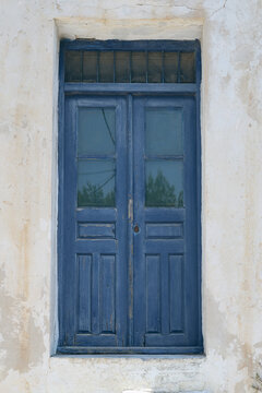 Tall weathered blue door with glass panes and transom window set in a cracked white wall, reflecting the aged elegance and coastal charm of traditional Greek island architecture.
