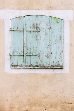 Faded mint‑green shutters framed in white against a beige wall reveal peeling paint and aged wood, showcasing the rustic charm and timeless texture of Mediterranean architecture.