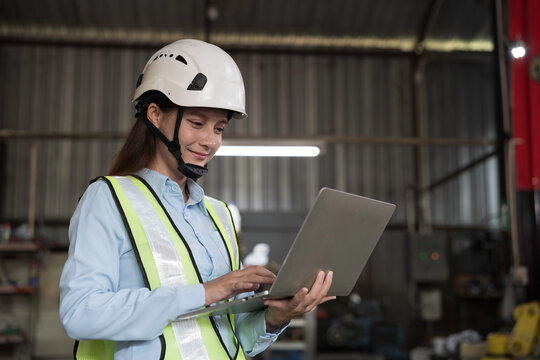 Woman engineer working with laptop computer at the industry factory area, wearing safety helmet, uniform checking and working in the factory