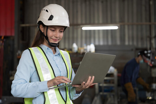 Woman engineer working with laptop computer at the industry factory area, wearing safety helmet, uniform checking and working in the factory