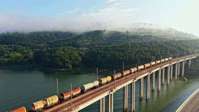 Long freight train with locomotive and connected wagons moves left to right across multi-span concrete viaduct above reflective river, with mist, sunlit hills, forested ridges, clear industrial