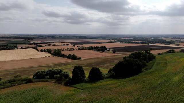 Green hill slopes toward open farmland fields. Clouds drift low over the distant horizon. Fields show brown crops and lush green grass. Trees line the land, adding natural structure