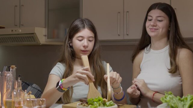 Two affectionate teenage sisters collaborating in the kitchen while laughing a lot and preparing a healthy salad together for a family dinner at home