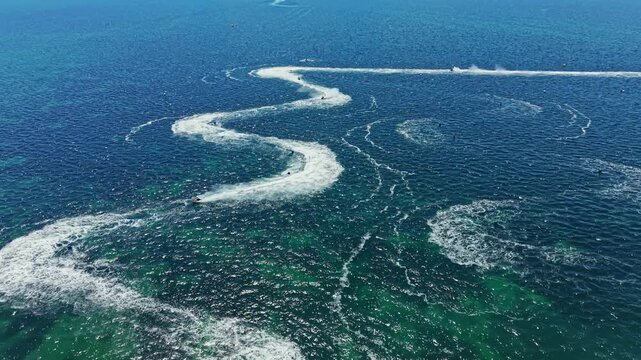 Racers maneuver jet skis in a winding pattern on clear water. The event takes place in a coastal area under bright daylight. Waves trace their paths behind them.