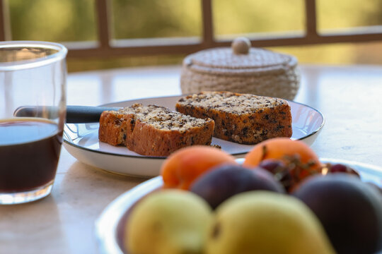 Two slices of homemade fruit loaf cake rest on a plate beside a glass of coffee. Fresh fruit in the foreground and a sunlit outdoor setting create a warm, inviting breakfast scene.