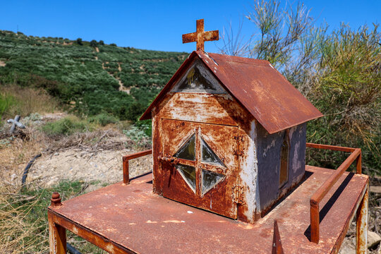 A small rusted devotional shrine with a cross sits on a metal stand in rural Crete, Greece. Olive-covered hills and native vegetation provide a striking Mediterranean backdrop.
