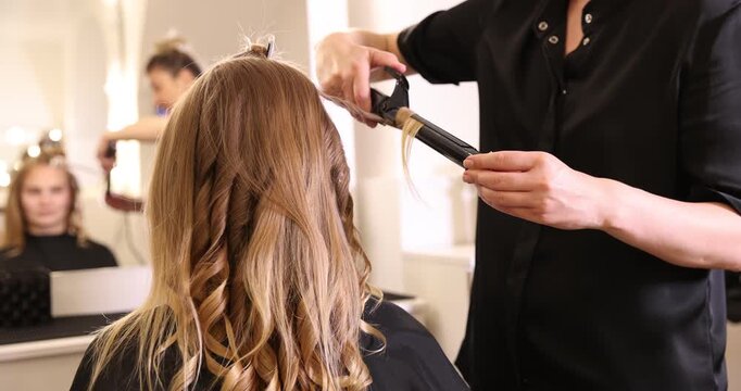 Hairdresser styling woman's hair with curling iron in beauty salon, closeup