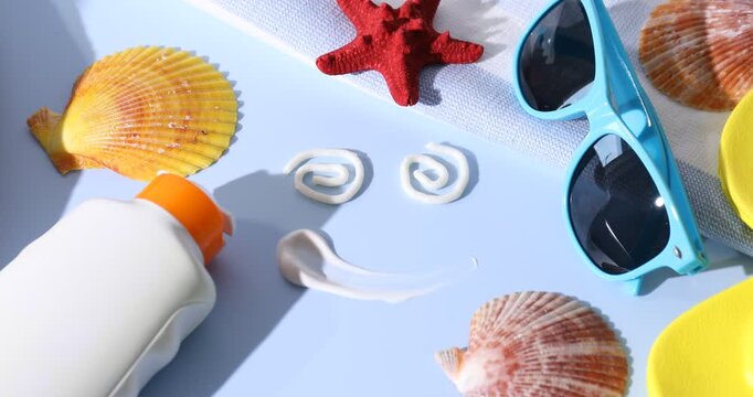 Smiling face made with sunscreen, bottle of sun protection product and beach accessories on light blue background, closeup. Camera moving in