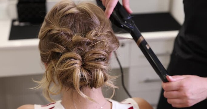 Hairdresser styling woman's hair with curling iron in beauty salon, closeup