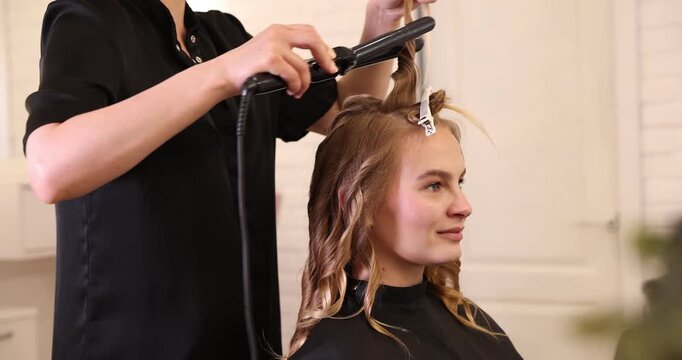 Hairdresser styling woman's hair with curling iron in beauty salon, closeup