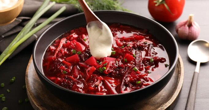 Adding sour cream into tasty borscht at black wooden table, closeup